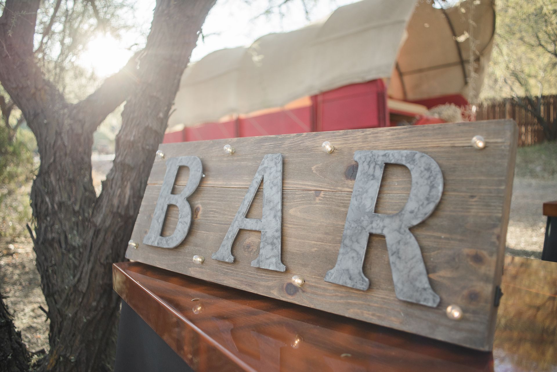 Country style wooden sign that reads BAR on table in sunny backyard with covered wagon in background
