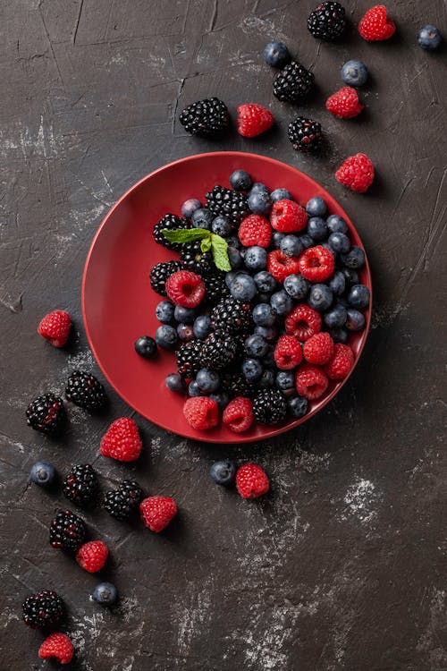 Top view of delicious blackberries and raspberries with blueberries on red round plate with more berries scattered around on dark grungy surface