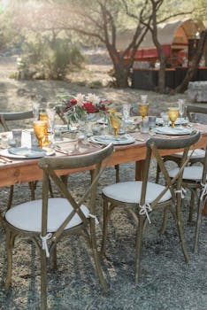 Elegant wooden table with wineglasses and flowers set for banquet in quiet backyard with no one around on sunny day with the Quote "Ideas pull the trigger, but instinct loads the gun." written on it and have average color value #7C776B