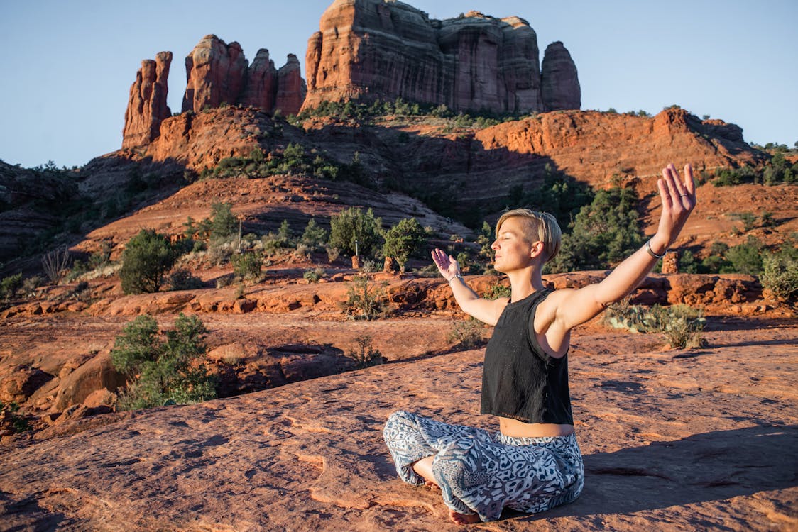 Free Young slim lady in summer top and hippie pants sitting with arms spread and eyes closed on rough ground while doing yoga near magnificent sunlit canyons Stock Photo