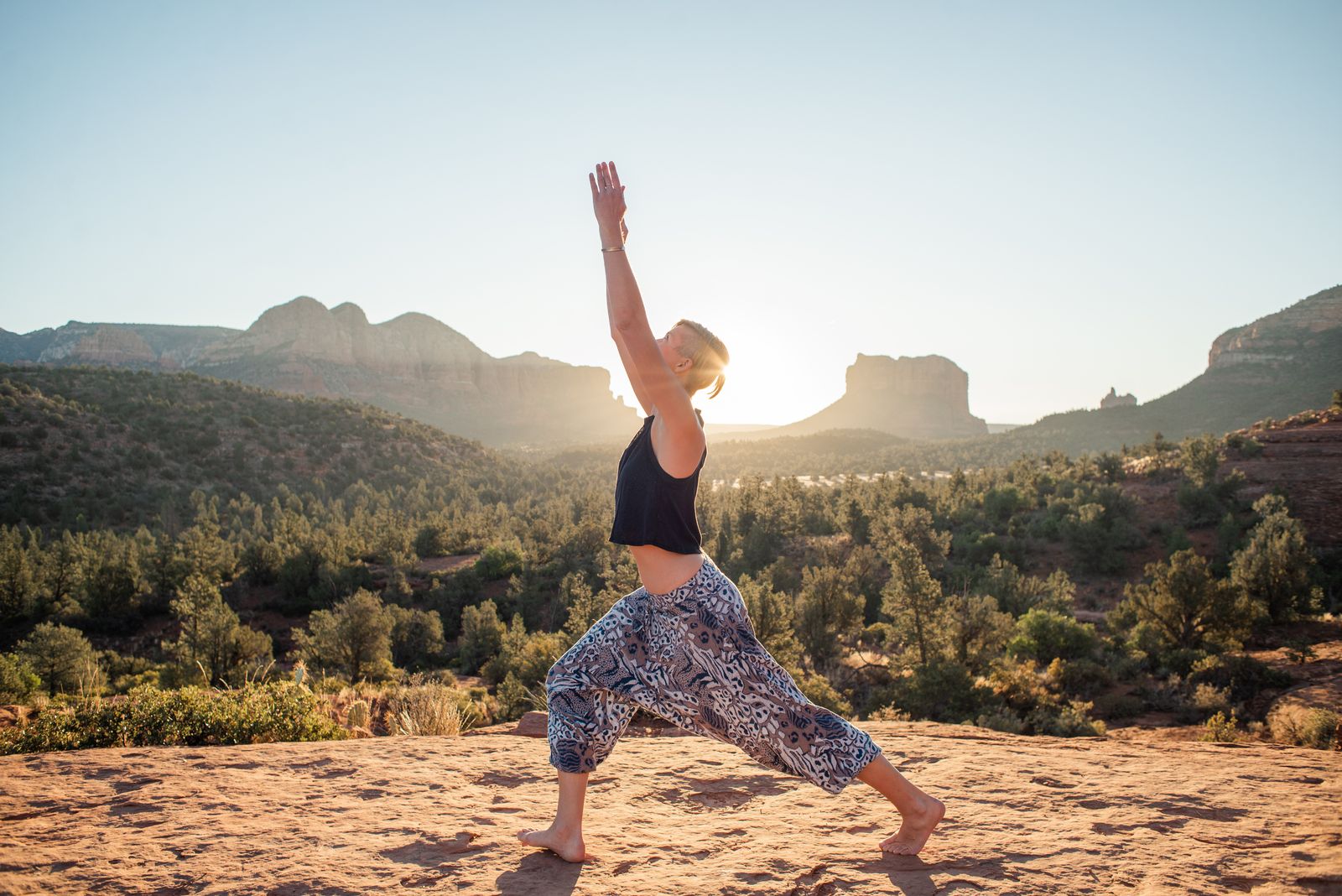 Woman practicing Anjaneyasana low lunge pose outdoors on a sunlit rocky landscape with arms raised overhead