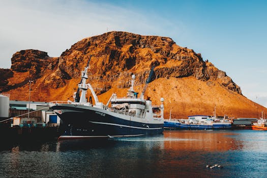 Colorful harbor scene in Vestmannaeyjar featuring ships against a rocky mountain backdrop under a clear blue sky.