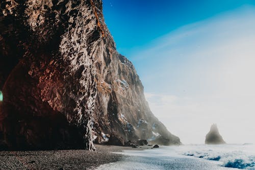 Vibrant blue sky with clouds above majestic rocky formation in sunlight on coast of wavy ocean