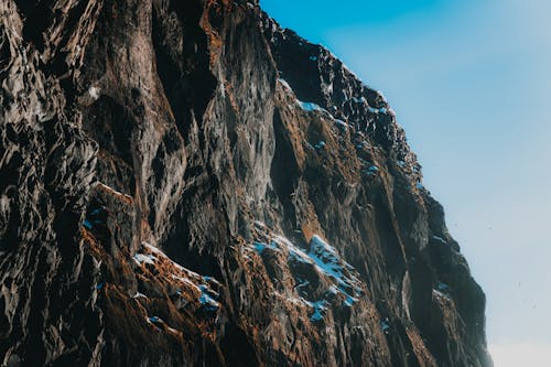 Picturesque view of light blue cloudless sky above majestic dark brown steep mountain covered with snow in daylight