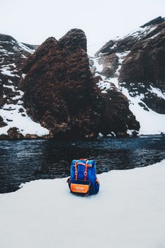 冬季，白雪皑皑的山脉旁，深蓝色的小背包挂在棕色肩带上，静静躺在黑暗河流的岸边