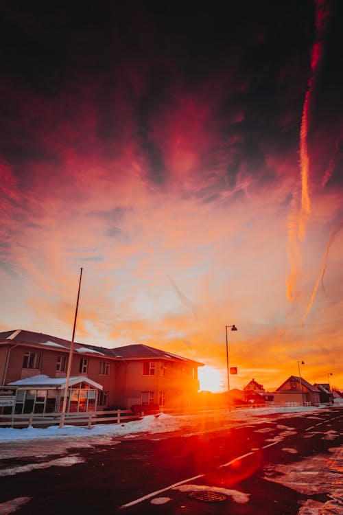 Amazing view of red sun shining on sundown sky over building located near snowy road against cloudy evening in winter