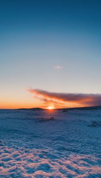 Picturesque view of mountain range in distance located near snowy area with sun on horizon against sundown sky in evening