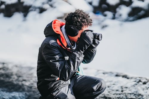 Side view of male photographer with curly hair in gloves squatting near snowy ground while taking pictures on camera of frozen scenery