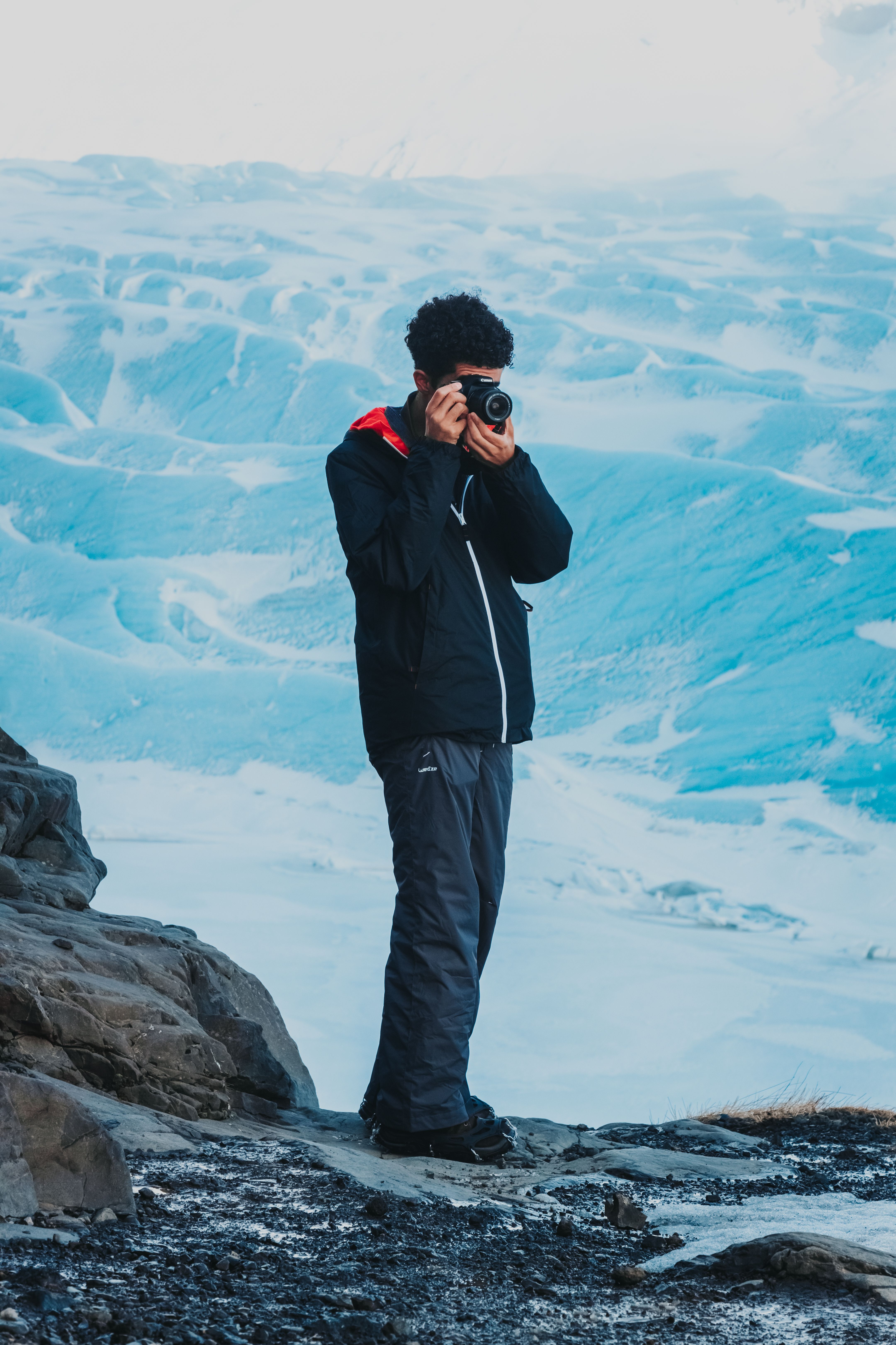 Faceless male camper standing on stony ground near snowy surface and taking photos of scenery in winter time in daytime