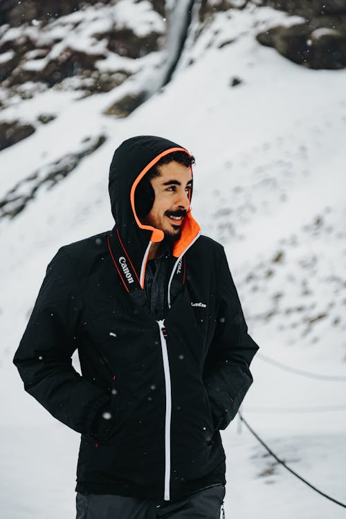 Optimistic male with mustache in hood standing with hands in pockets while admiring view of snowy nature during winter weekend