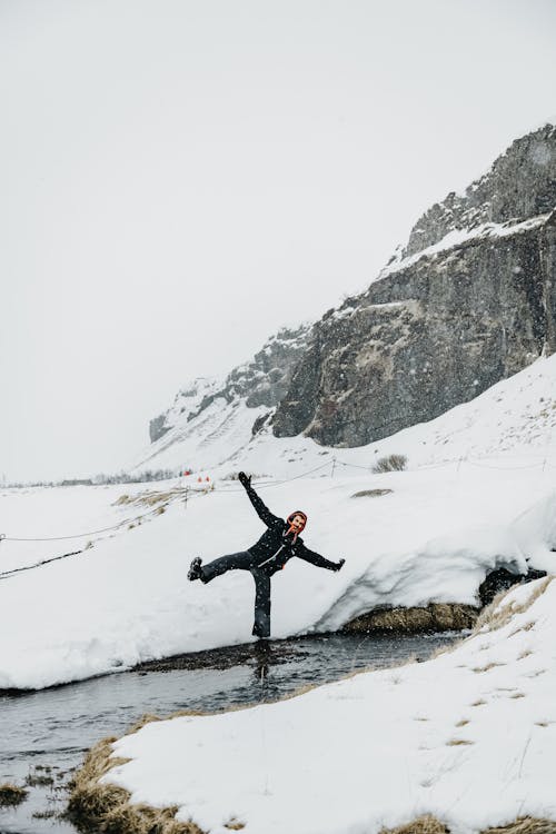 Full body of anonymous traveler standing on one leg in mountain river near snowy hills against gray sky during vacation