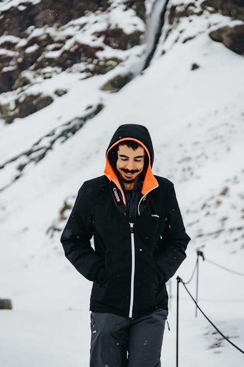 Cheerful male traveler in warm outerwear standing near rocky cliff covered with snow during trip through mointainous terrain in winter