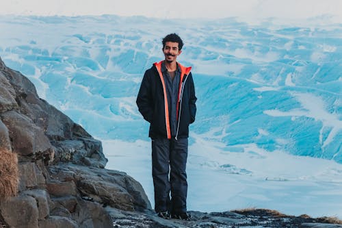 Full body cheerful man in winter outerwear smiling at camera while standing on rocky range in snowy valley