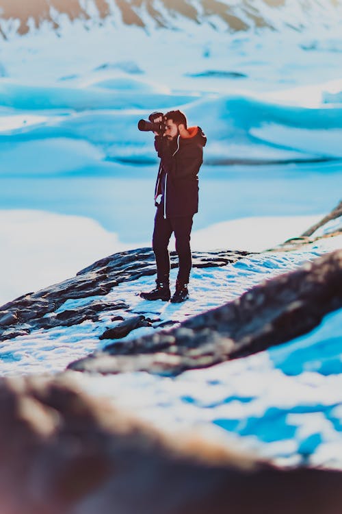 Full length of ethnic man in winter clothes taking photo of snowy amazing landscape