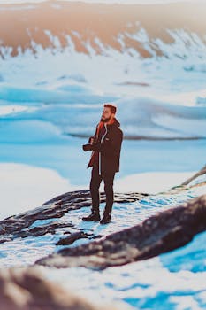 A man in warm clothing photographs a breathtaking snowy mountain landscape.