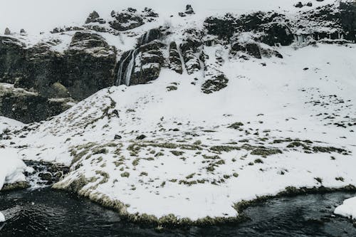 Peaceful water flowing alongside cold coast covered with snow against rocky cliffs in gloomy day