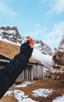 Faceless person in warm clothes holding transparent clear icicle in hand standing against aged cabin in cold mountains