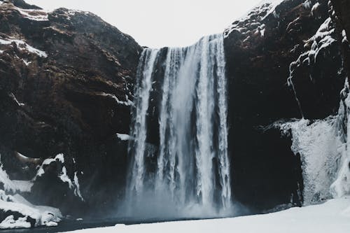 Rapid foamy cascade in rough mounts near pond and snowy terrain under sky in winter