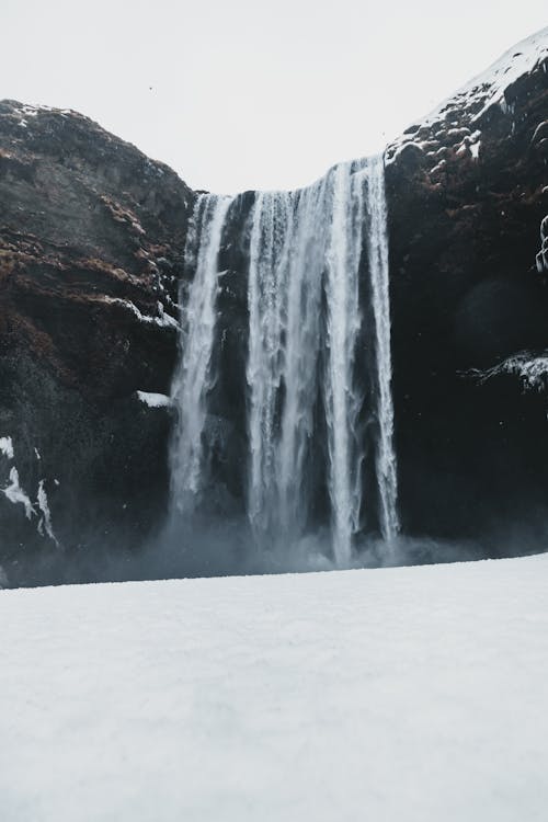 Scenery view of fast foamy waterfall in bristly mounts near snowy surface under sky