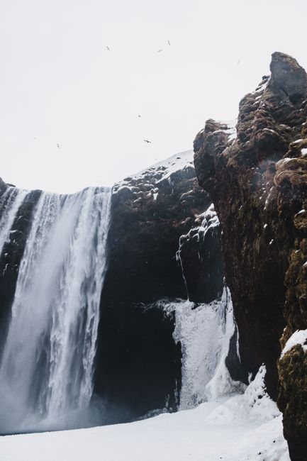 From below of fast waterfall in rough mounts near snowy terrain under white sky with flying birds