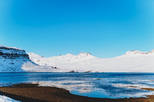 Serene winter landscape of snow-covered mountains and a clear blue lake under a bright sky.