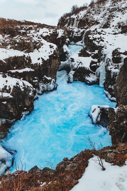 From above picturesque view of bright cascade and pond in rough mounts with snow under cloudy sky