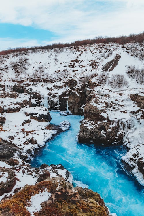 From above of narrow blue creek between rough mounts covered with snow under sky in winter