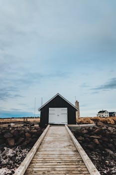 在多云的天空下，迷人的冬日场景里，岩石海岸边有一座乡村房屋，旁边是一条木制步道