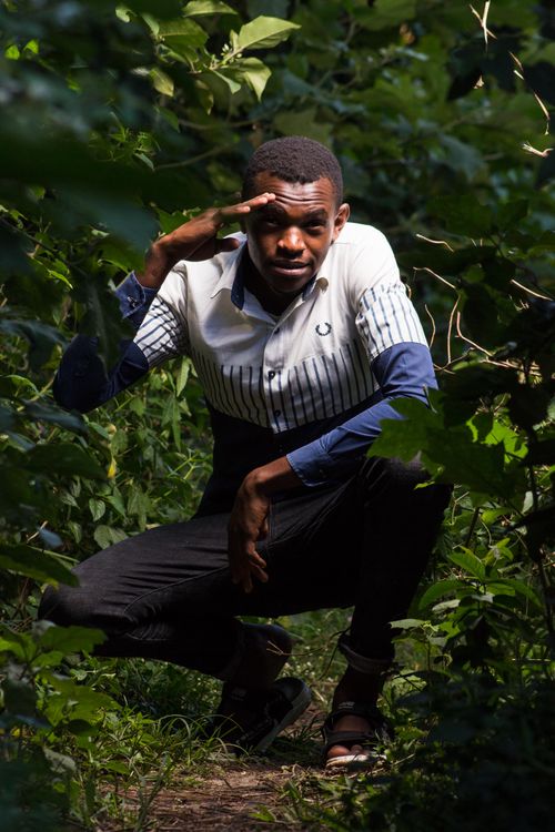 Free Young man squatting in a green forest, saluting with one hand, wearing a striped shirt and black pants. Stock Photo