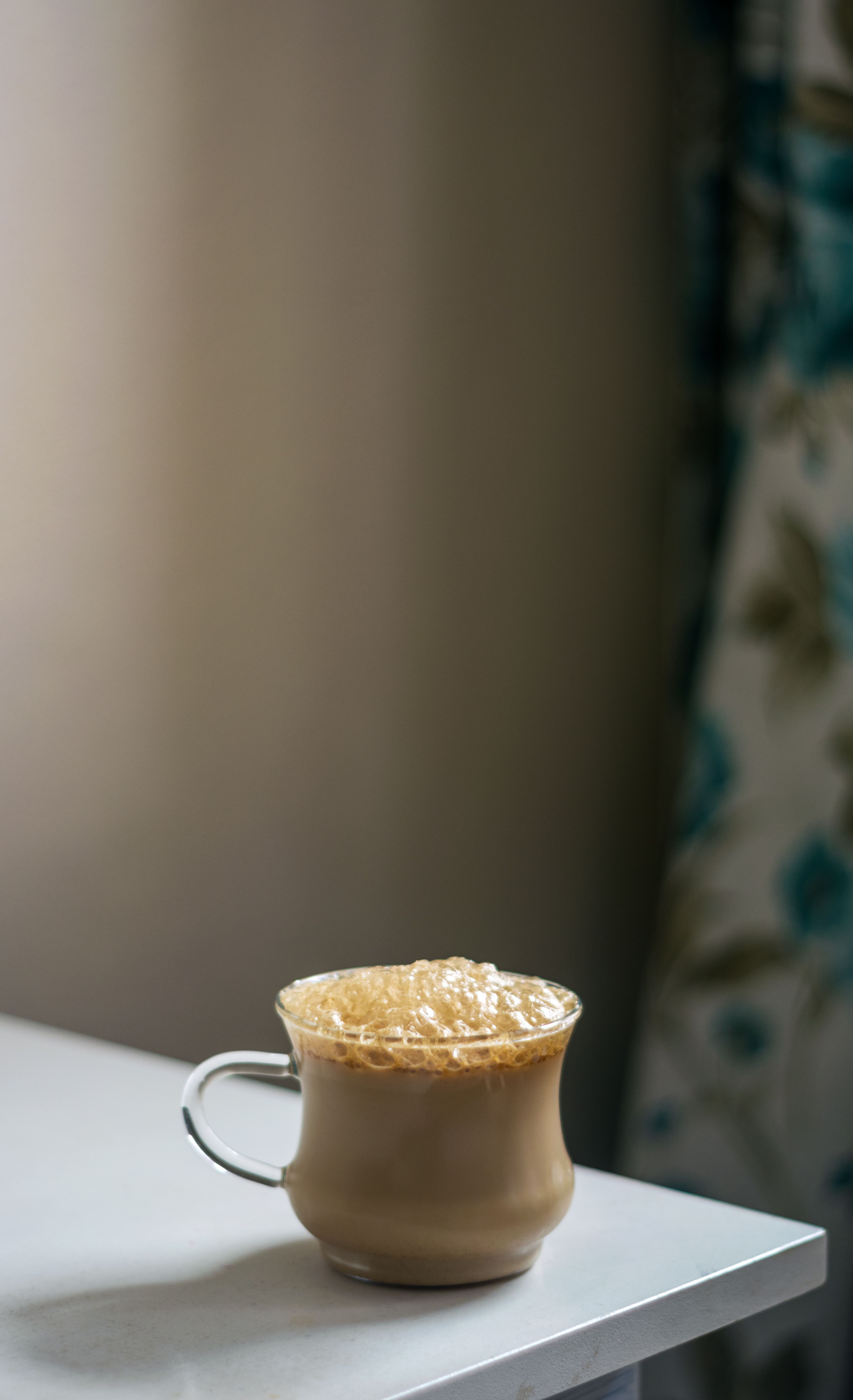 A glass cup of cappuccino with rich foam sits on a white table, creating a warm and inviting atmosphere.