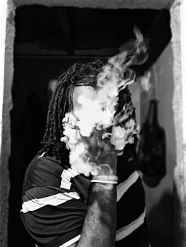 A faceless man vaping in an urban setting, surrounded by smoke in a black and white photograph.