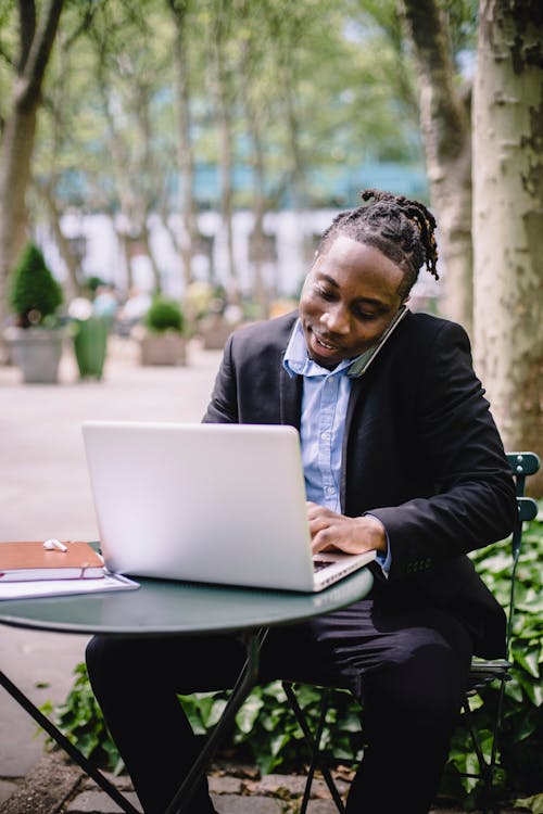 Free Happy African American businesswoman in formal clothes having conversation via mobile phone while working on netbook in summer park Stock Photo