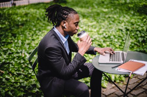 Free Side view contemplative black businessman in formal outfit surfing modern netbook while enjoying hot drink and listening to music via earbuds in sunny park Stock Photo