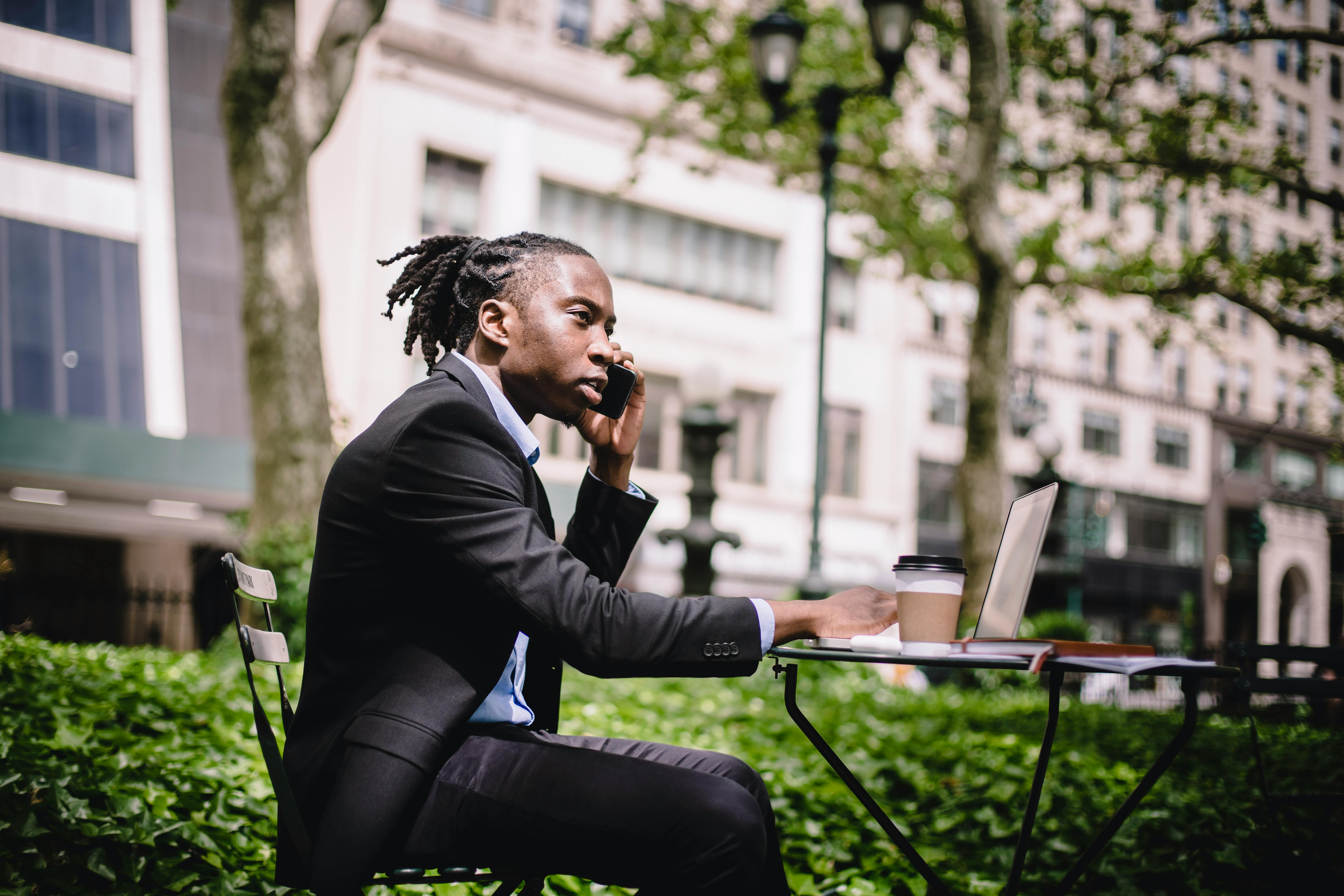 thoughtful black businessman with laptop talking on smartphone in garden