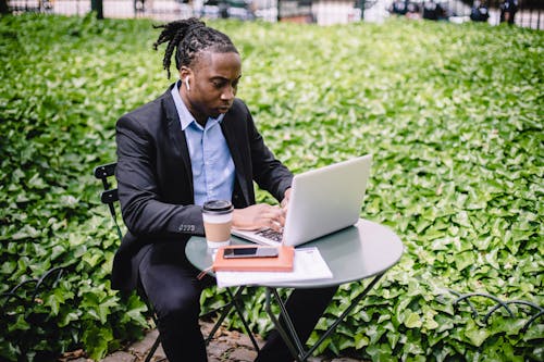 Concentrated African American businesswoman in earbuds wearing formal black suit sitting at table in city garden and typing on netbook