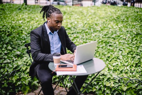 Free Concentrated African American businesswoman in earbuds wearing formal black suit sitting at table in city garden and typing on netbook Stock Photo