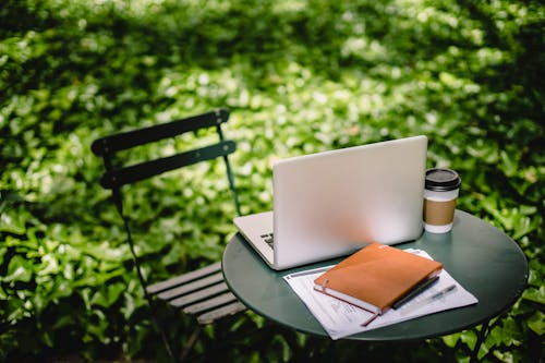 Free Modern netbook with notebook placed on small round table near paper cup with hot drink in sunny green park Stock Photo