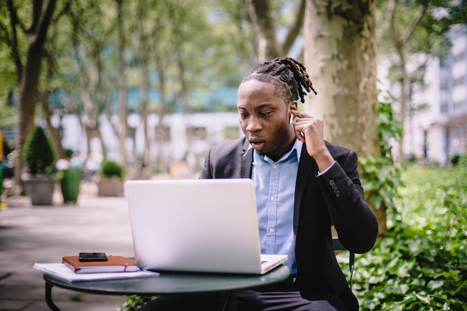 Concentrated young black male with dreadlocks in formal wear having video conversation on laptop using TWS earphones while sitting in outdoor cafe
