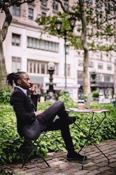 Full body of young African American male entrepreneur in suit talking on mobile phone sitting at table in outdoor cafe