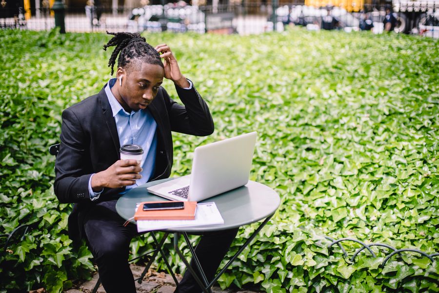 Focused young African American male freelancer with dreadlocks in formal wear drinking coffee in street cafe and scratching head while reading report on laptop