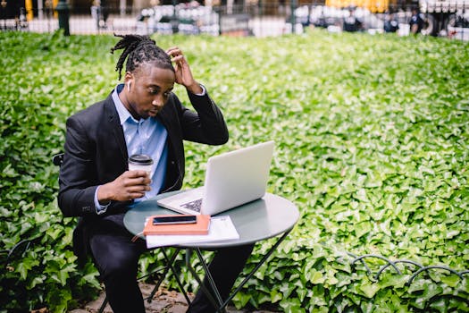 Focused young African American male freelancer with dreadlocks in formal wear drinking coffee in street cafe and scratching head while reading report on laptop