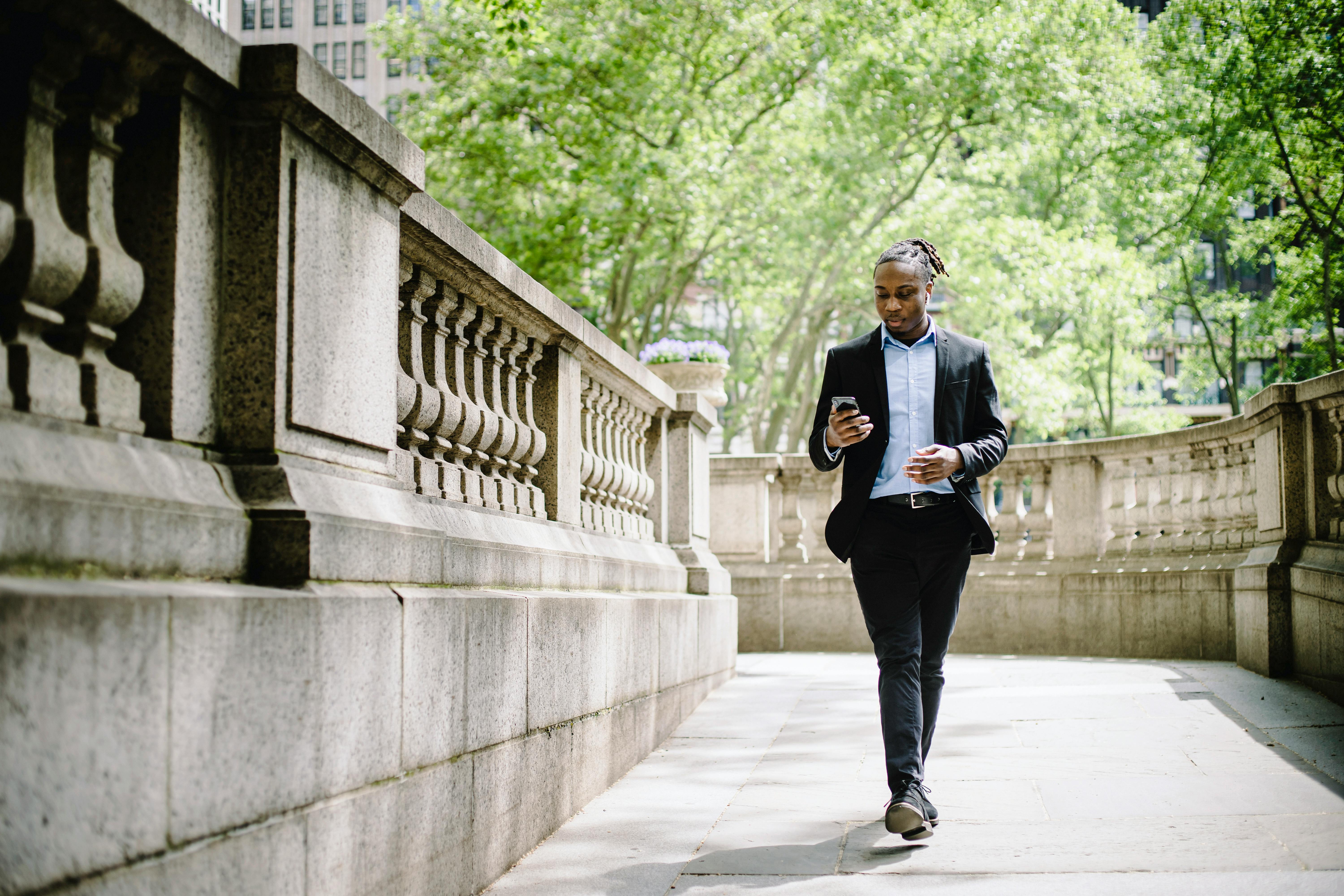 concentrated black businessman walking in city park and messaging on smartphone