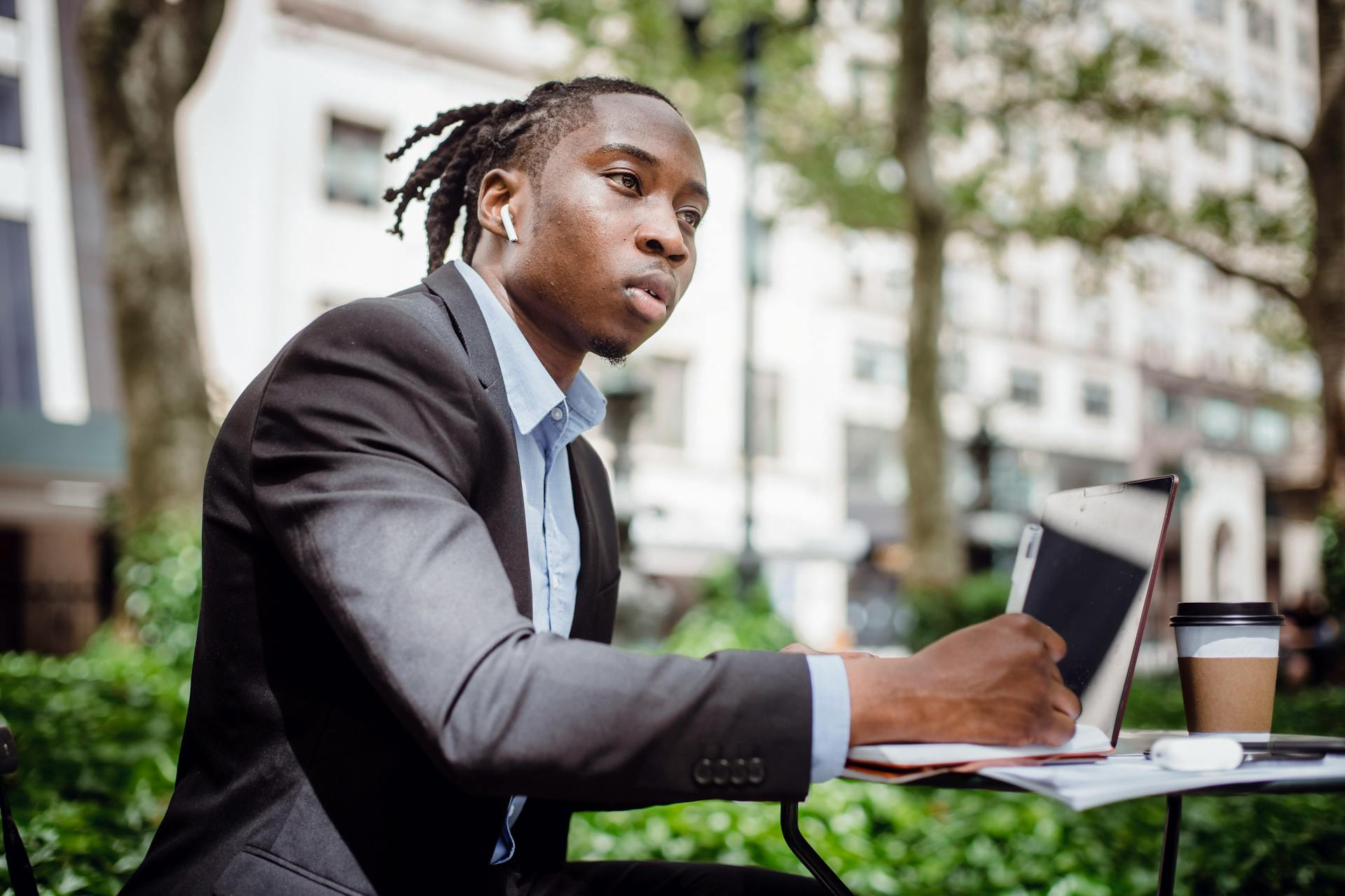 Side view of young black male writer in suit and true wireless earphones looking away and thinking while taking notes in planner sitting in street cafe with laptop and coffee