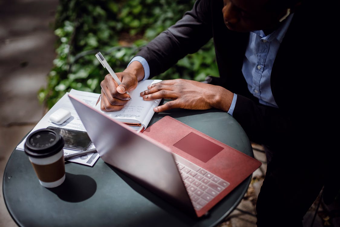 From above of crop black male journalist in formal wear writing article while sitting at table with red laptop and cup of takeaway coffee