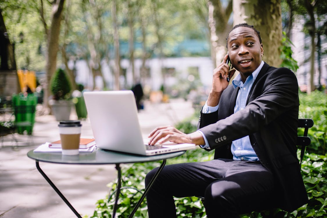 Free Cheerful young African American male entrepreneur having phone call while working remotely on laptop in street cafe on sunny day Stock Photo