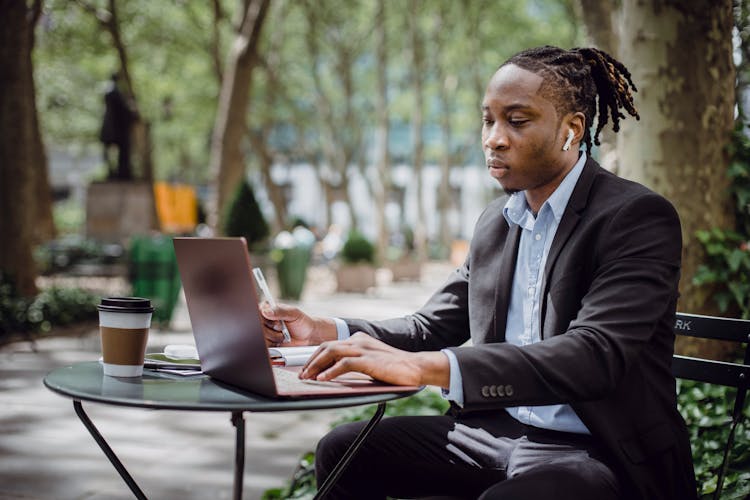 Busy Young African American Man With Dreadlocks Working On Laptop In Street Cafe