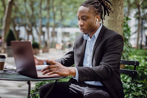 Free Serious young African American male with dreadlocks in suit and TWS earphones sitting in street cafe and working remotely with laptop Stock Photo