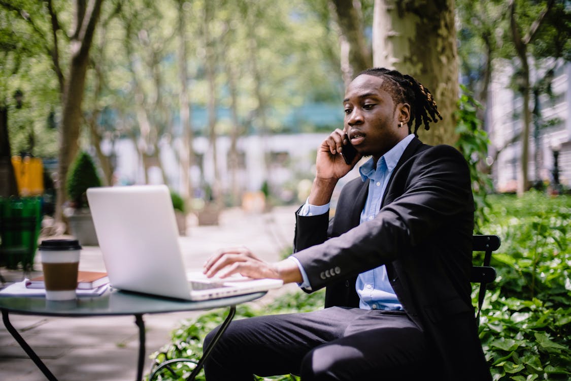 Free Serious stylish young African American male manager in formal wear browsing laptop while having phone call in outdoor cafe Stock Photo