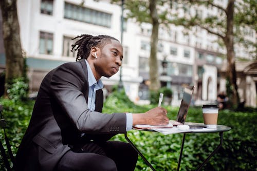 Side view of concentrated young black male writer listening to music with true wireless earbuds and looking away while creating new article in street cafe