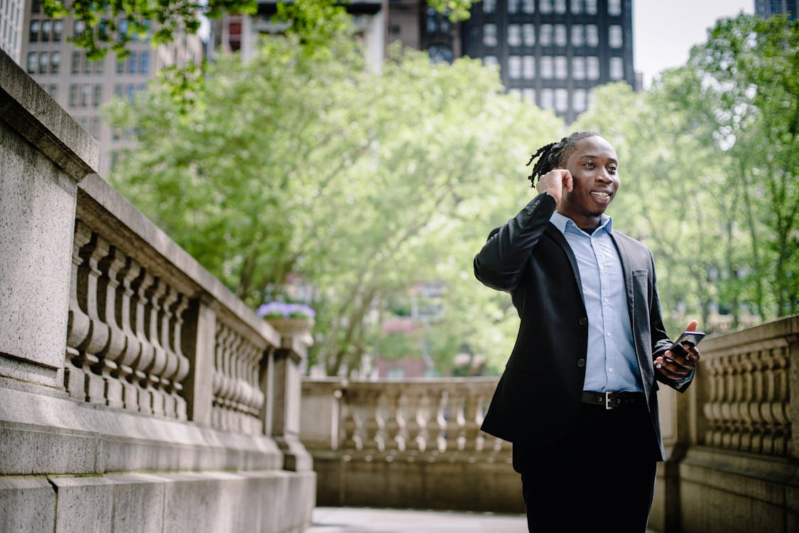 Happy young African American male manager in formal suit with smartphone answering call with wireless earphones against modern buildings in business district